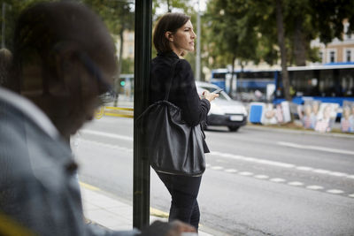 Businesswoman looking away while standing at bus stop with commuter in city