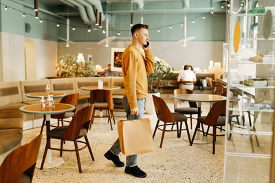 A young man in casual clothes with bags in his hands after shopping stands alone and talks in a cafe