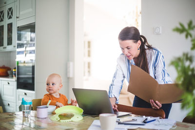 Female professional holding file while using laptop by daughter on dining table at home office