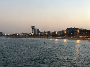 Sea by illuminated city against sky at dusk