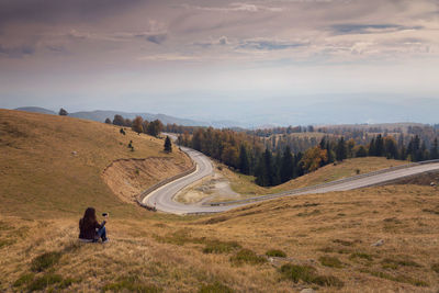 Rear view of woman holding mobile phone while sitting on field by road against sky