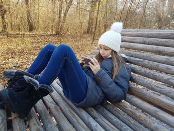 Full length of girl using mobile phone while lying on wood in forest during winter