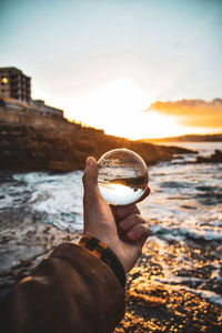 Person holding glass of water at beach against sky during sunset