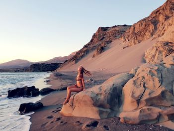 Woman in bikini sitting on rock at beach against sky