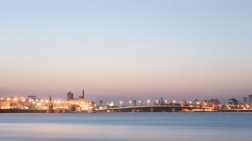 Illuminated buildings in front of river against sky at dusk in city