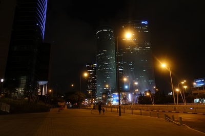 Illuminated street amidst buildings against sky at night