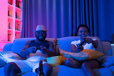 Portrait of couple holding video game remote control while sitting on sofa in living room at home