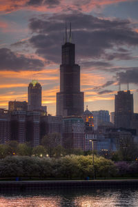 Modern buildings by river against sky during sunset
