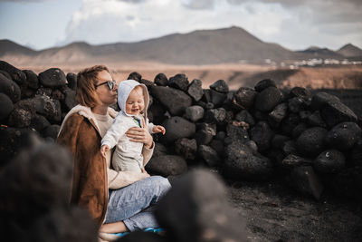 Mother enjoying winter vacations playing with his infant baby boy son on black sandy volcanic beach