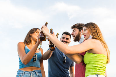 Cheerful friends toasting beer bottles while standing against sky