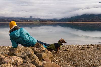 Rear view of unrecognizable woman resting on the rock with her dachshund teckel dog near to the lake