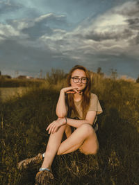 Young woman sitting on grass at field against sky