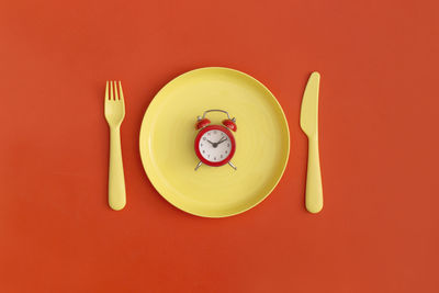 Red clock on the yellow plate with fork and spoon on a red background. top view. flat lay.