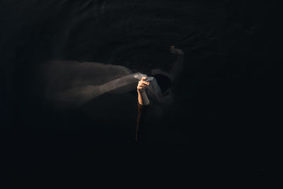 From above side view of unrecognizable female traveler covering face with textile while standing with raised arms in water at dusk