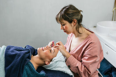 Side view of female cosmetologist plucking eyebrows on face of woman client lying on table in beauty salon
