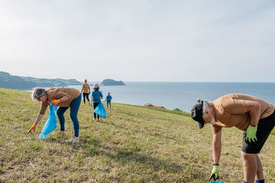 Eco activists of different ages in sportswear with blue bin bags collecting trash on green grassy hillside near calm blue sea on sunny day