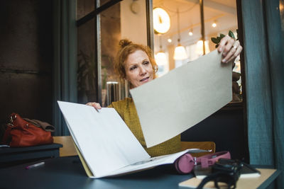 Confident mature female illustrator holding papers at desk in creative office