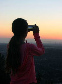 Rear view of girl photographing sunset from mobile phone