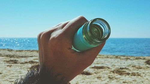 Close-up of hand holding bottle at beach against clear blue sky