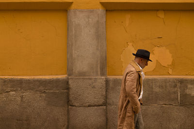 Adult man in hat and coat walking in front of yellow wall on street during sand storm day. madrid.