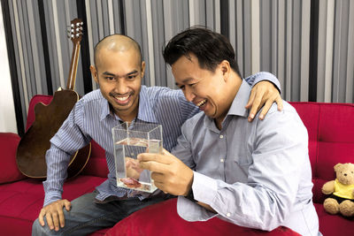 Happy friends holding glass container with water while sitting on sofa at home
