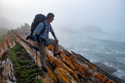 Side view of man with backpack and trekking pole on orange sea rocks looking at crashing waves 