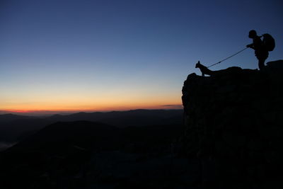 Silhouette man with animal on cliff against sky during sunset