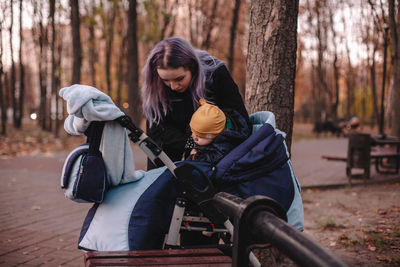 Young mother putting baby son in baby stroller in park during autumn