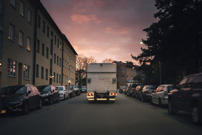 Cars on road by buildings against sky during sunset