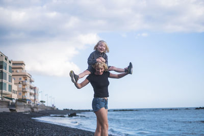 Happy woman carrying son on shoulder at beach against sky