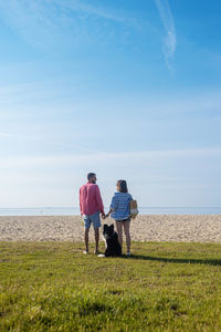 Back view of couple standing and holding hands near their dog on beach