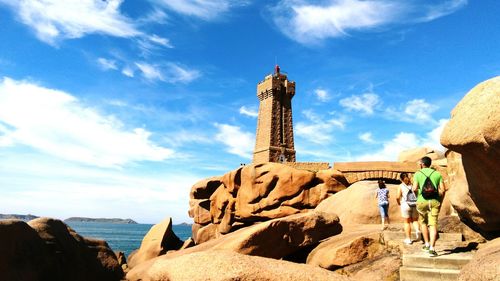 Friends walking towards lighthouse by sea against cloudy sky during sunny day