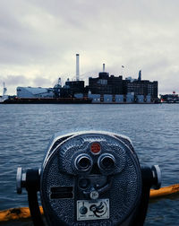 Close-up of coin-operated binoculars by river against buildings and sky