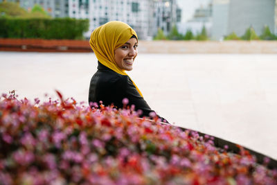 Side view of cheerful muslim female in headscarf sitting on bench near flowerbed in street and looking at camera