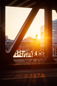Woman standing by bicycle on bridge in city during sunset