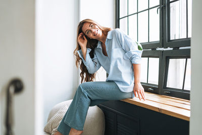 Portrait of young smiling woman with long hair sitting on window sill in modern office