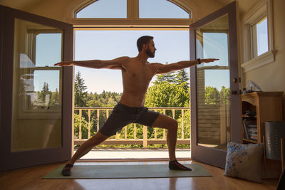 Man doing warrior yoga pose in front of open window doors to forest view.