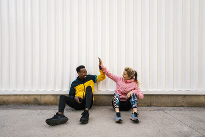 Male and female athlete doing high-five while sitting on sidewalk against wall