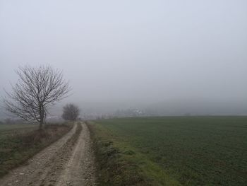 Dirt road amidst field against sky during foggy weather