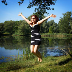 Full length portrait of woman in mid-air with arms outstretched at lakeshore against sky during sunny day