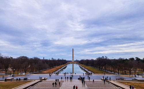 People in city and washington monument against cloudy sky