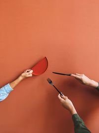 Cropped hands of female friends holding plate and eating utensils against orange wall