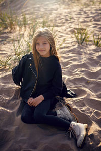 Portrait of a girl with long hair sitting on a sandy beach in a black leather jacket