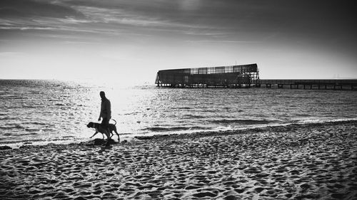 Silhouette man with dog walking on sandy beach during sunny day