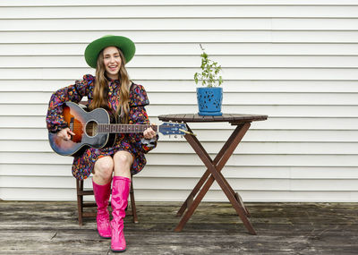 A woman sits alone at a wooden table on deck playing acoustic guitar