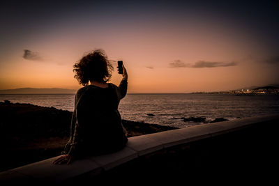 Rear view of woman taking a selfie against sky at sunset