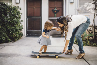 Daughter skateboarding with help of smiling mother on footpath