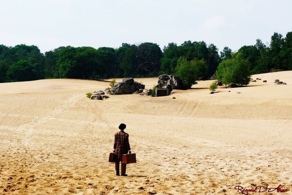 REAR VIEW OF A MAN STANDING ON SANDY LANDSCAPE