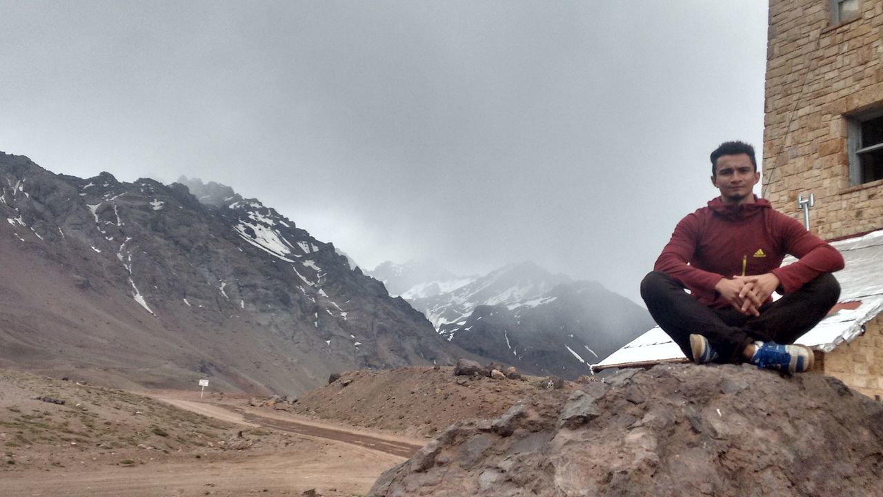 lifestyles, leisure activity, mountain, full length, young adult, casual clothing, person, young men, winter, warm clothing, sky, sitting, rock - object, cold temperature, standing, snow, mountain range