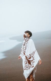 Young man wrapped in fabric standing at beach during foggy weather